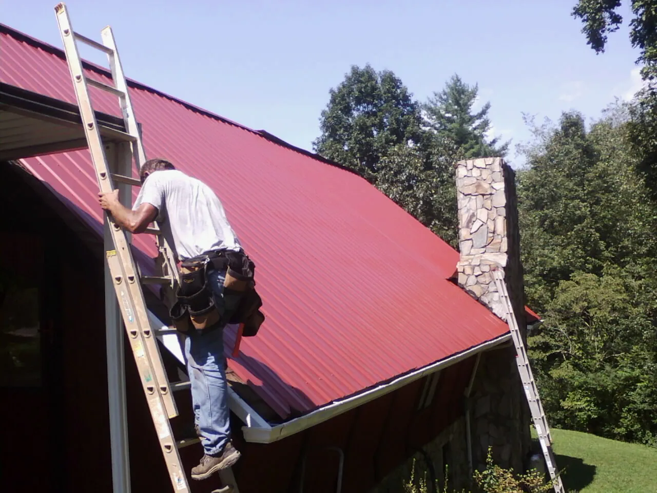 Roofer on a ladder beside a red metal roof and stone chimney — Soffit & Fascia Repair by Roofing Brothers in Stevenson